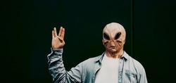 Alien mask people against a black background doing hallo gesture with hand. Portrait of extraterrestrial with human casual clothes. Banner header for immigration concept and invasion