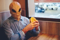 Alien with human clothes eating hamburger sandwich inside a fast food sitting at the table alone. Extraterrestrial and immigration invasion concept. Junk food nutrition unhealthy