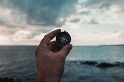 Pov of man hand holding navigational travel compass to find direction and next destination place. Concept of travel people and exploring. Beach and ocean with sky light in background