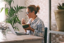 Side view of adult young woman smiling and eating in fron tof a laptop at the desk at home. Alternative office lifestyle female people working in video call conference during lunch time. Modern job