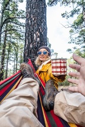 Cheerful woman with cup of coffe enjoy the nature relax outdoor leisure activity with man laying on the hammock - happy people in forest woods freedom lifestyle together - travel and environment