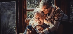 Happy senior old couple use mobile phone at home together to video call parents. Winter season indoor leisure activity for retired mature people. Woman hug mature man on the chair. Love and technology