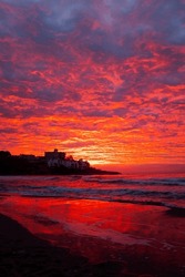 Red sunset at the beach with town in silhouette and dramatic sky with clouds. Amazing sunlight sunrise seascape with waves and houses in background. Concept of travel destination without people