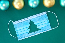 Blue medical protective mask with cut-out silhouette of Christmas tree on green background. With Christmas balls near mask. Christmas and Happy New Year concept, coronavirus concept. Soft focus
