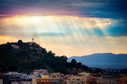 Beam of light through the clouds over a cross on top of the hill. Rays of light shining through clouds. Celestial entity over a hill with a crucifix that protects the city. Cross silhouette.