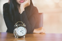 wasting time concept with Asian business woman feeling tired and bored waiting for someone coming late at work and looking at alarm clock on desk  
