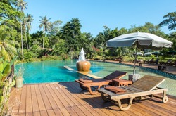 Relax in The Luxury Hotel on The Island of Thailand. Couple of Wooden Chair on Terrace under The Umbrella by The Beautiful Luxury Swimming Pool surrounded by Various Type of Palm Trees in The Garden.