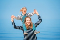Close-up of a happy family on the beach on a sunny day. Daughter sits on mother's shoulders, arms outstretched on the beach. Smiling mother and daughter playing on the beach