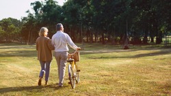 The behind of Caucasian elderly couples walking with a bicycle in the natural autumn sunlight garden feel cherish and love, concept elderly love, warm family, happy retirement, retirement lifestyle.