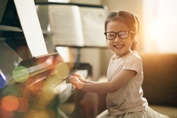 Asian toddler girl look at camera with smiling while playing piano, Happy back to school concept, Early childhood music education, leisure activity, side view girl kid sitting with piano.