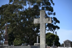 Weathered stone cross in a cemetery on a sunny day, with graves, a large tree, and blue sky in the background