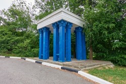 The pavilion of the old bus stop and the forest next to the mountain road. Antique blue carved columns and white roof. There is a black and white curb along the road. Selective focus.