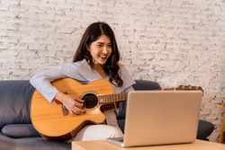 Young Asian woman practicing and learning how to play guitar on laptop computer monitor. Female guitarist watching online tutorial.