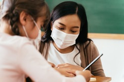 Close-up of Asian female teacher wearing a face mask in school building tutoring a primary student girl. Elementary pupil is writing and learning in classroom. Covid-19 school reopen concept