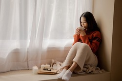Young beautiful Asian woman holding cup of coffee and marshmallows on top, sitting at home and looking out the window. Happy girl drinking chocolate in sweater in cold weather winter