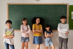 Happy multiethnic children in casual clothes crossing arms and smiling while standing near chalkboard during lesson at primary school