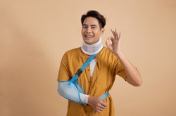 Handsome young asian man with broken arm in soft splint suffering a sore arm showing ok sign isolated on beige background, accident insurance concept.