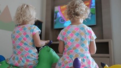 Children watch cartoons on TV in the playroom. Two blond girls sit on soft pillows in front of the TV and watch children's cartoons. The concept of teaching children TV from an early age.