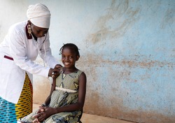 Black pediatrician with a doctor's suit is placing a stethoscope on the chest of a smiling little girl with typical African braids during routine respiratory check-up outdoors a rural hospital setting