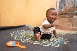 Cute little African baby boy sitting on a carpet with toys around him pointing at someone with a big confident smile on his face; symbol of childish innocence and trust