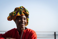 Portrait of a radiant African beauty wearing a red caftan and a colorful headscarf with a traditional pattern sitting casually by the river