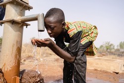 Thirsty young black African boy leans forward to drink fresh and clean water with his bare hands from a tap; water scarcity concept