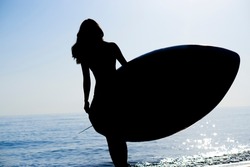 The dark silhouette of a slender girl surfer walks along the ocean and holds a board for  SUP water sports in the rays of the sunset.