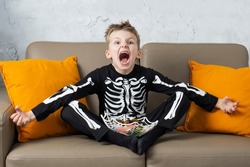 Happy shocked crazy shouts halloween boy having fun at home eat scary candy and looking at camera wearing Halloween costume skeleton on home background.