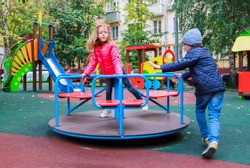 boy rolls girl in pink jacket on carousel in yard at playground outdoors