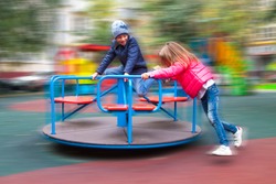 girl in pink jacket rolls boy on carousel in yard at playground outdoors