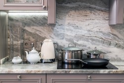custom designed kitchen, with marble looking quartz countertop and backsplash. cream electric kettle with porcelain tea accessories on granite countertop next to ceramic hob with steel pots and pan