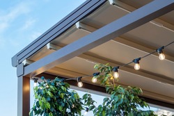canvas awning with metal frame and luminous garland of light bulbs against blue sunny sky. modern shading for house