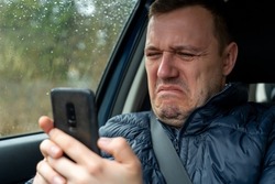 Sad millennial guy looks at phone screen in surprise, sits in passenger seat. Disgusted and overwhelmed man stares at screen of smartphone, needs to pay debts immediately. Gray rainy day