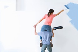 Woman is applying blue paint to the wall using a paint-roller while sitting on her husband's shoulder. 