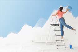 Woman standing on a ladder is applying blue paint to the wall in her house using a paint-roller with her back to the camera.
