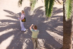 High angle view of a man with digital camera taking a picture of his wife on a sidewalk under the shade of a palm tree.