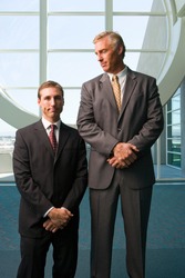Vertical three quarter front view of two businessmen standing besides each other with the taller one looking at the shorter in office lobby.