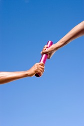 Close up of a female athlete passing a relay baton to her teammate on a bright, sunny day at an athletics competition