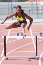 Young African female runner jumping over a hurdle at an athletics event out at the track on a bright, sunny day