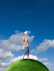 A happy boy throwing paper airplane from the top of a grassy globe against the backdrop of a bright, blue sky.