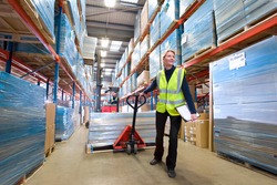 A wide shot of a female warehouse manager holding a clipboard and pulling a pallet truck.