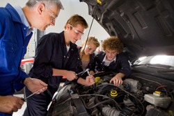 A side view of a young auto mechanics student repairing a car while being observed by his trainer and other students in an automotive training school