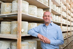 A medium shot of a owner standing in a cellar and smiling with aged cheddar cheese wheels in background.