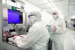 A scientist in a clean suit working on a computer next to a colleague walking in with motion blur in the silicon wafer manufacturing room