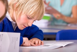 A tight closeup of a school boy in a classroom under selective focus intently reading a textbook while pointing and sliding his finger over each word