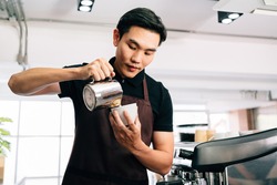 A young Asian barista man in an apron dress intentionally poured hot milk into a hot espresso black coffee for making latte art.