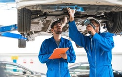 Mechanic in blue workwear uniform inspects the car bottom with his assistant. Automobile repairing service, Professional occupation teamwork. Vehicle maintenance. Focus on mechanics.