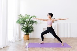 Young smiling attractive sporty Asian woman practicing yoga, doing Virabhadrasana 2 exercise, meditating in Warrior Two yoga pose, indoor working out at home, wearing sportswear. Full length photo.