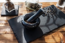 Black marble mortar with a pestle full of ground seasonings. Cutting slate board with a mixture of peppers and a glass bottle with spices. Kitchen utensils on a wooden table background.