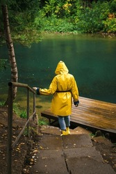 Woman in a yellow raincoat and rubber boots descends the stairs to the pier by the lake in a green wet forest during the rain. The concept of travel and tourism.